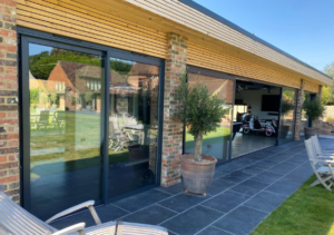 Long single-storey building with timber overhang supported by brick pillars and large sliding glass doors; dark slate patio with potted trees and lounge chairs, with view of red brick outbuilding and green lawn; part of Glaze Ltd project